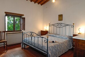 A bedroom with a metal bed frame, a wooden chair, a radiator, a window with a view of greenery, and bedside lamps.