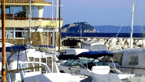 A marina with boats docked, a building with a sign reading "PORT", and a rocky coastline with a castle-like structure in the background. - Les Voiles d'Azur (La Londe-les-Maures)