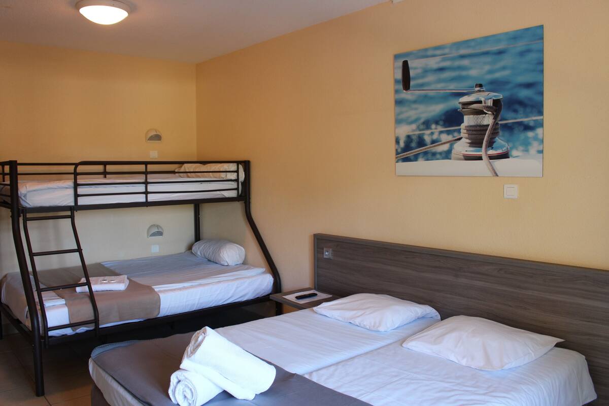Una habitación con literas y una cama individual, una pequeña mesa con un control remoto y un cuadro de un barco en la pared.