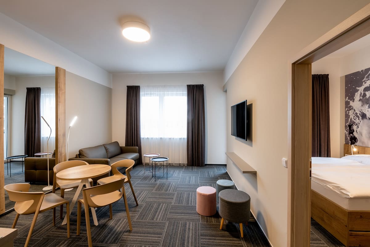 A hotel room with a bed, a sofa, a dining table with chairs, a TV, and a large window with curtains.