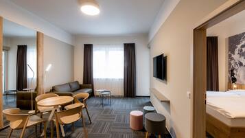 A hotel room with a bed, a sofa, a dining table with chairs, a TV, and a large window with curtains.