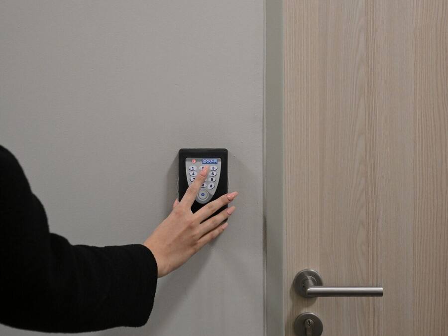 A hand pressing a doorbell button on a gray door with a yellow sign above it reading "Apartment 13".