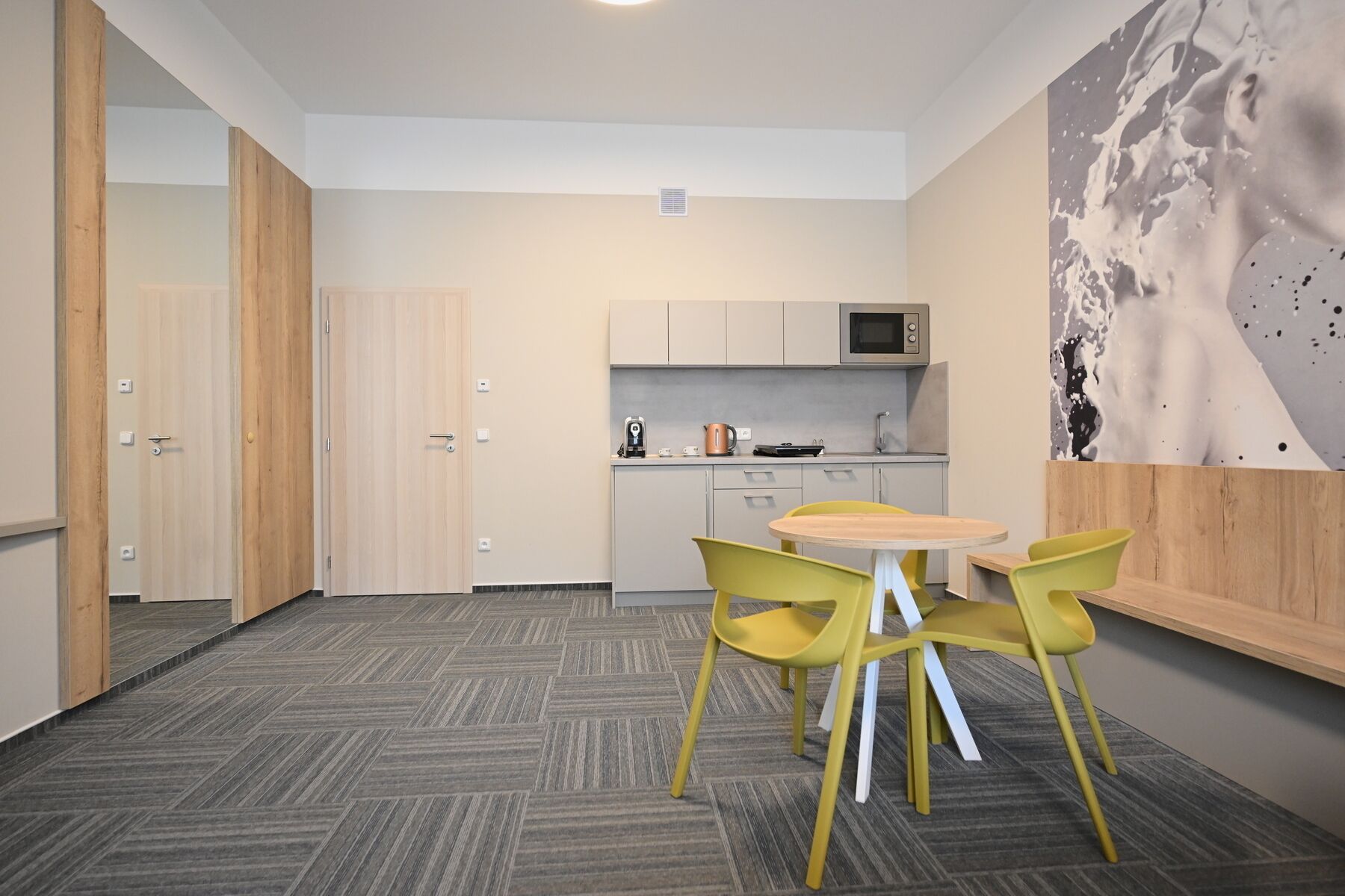 A compact kitchen with a microwave, kettle, and coffee maker. There is a small dining area with a round table and yellow chairs. The room has a herringbone-patterned carpet and wooden doors.