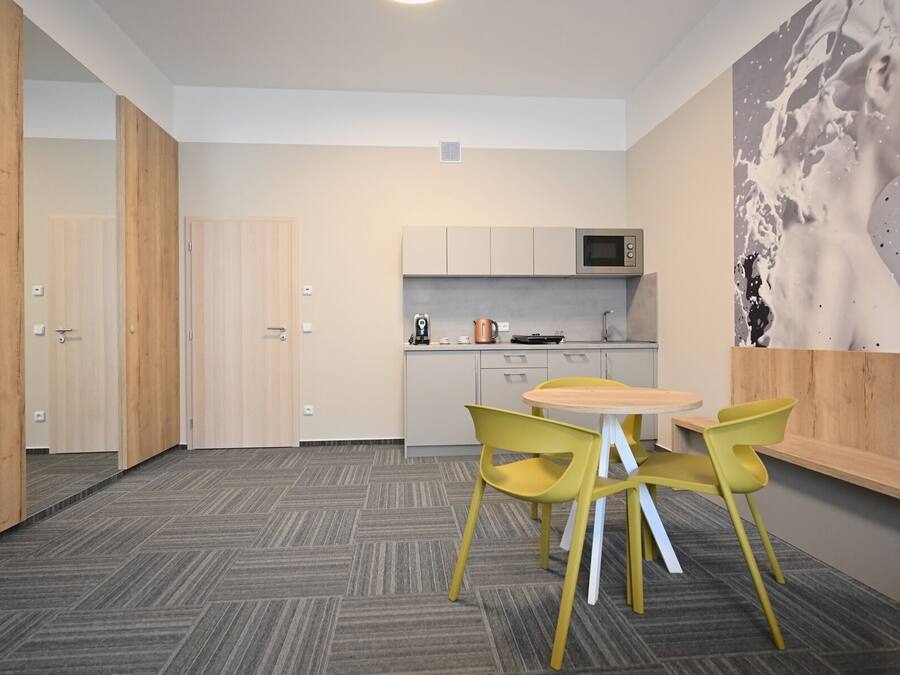 A compact kitchen with a microwave, kettle, and coffee maker. There is a small dining area with a round table and yellow chairs. The room has a herringbone-patterned carpet and wooden doors.