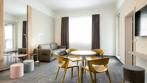 Une chambre d’hôtel moderne avec une table ronde, des chaises jaunes, un canapé et un téléviseur à écran plat.