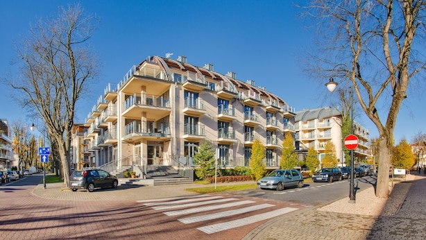 A multi-story residential building with balconies, a pedestrian crossing, and parked cars.