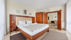 A hotel room with a large bed, wooden headboard, a bedside table, a small drawer, a brown rug, and a door leading to another room.