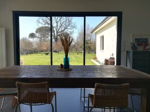 Un coin repas avec une grande table et deux chaises en osier, un vase contenant des plantes séchées et une vue sur un jardin, visible à travers de grandes fenêtres.
