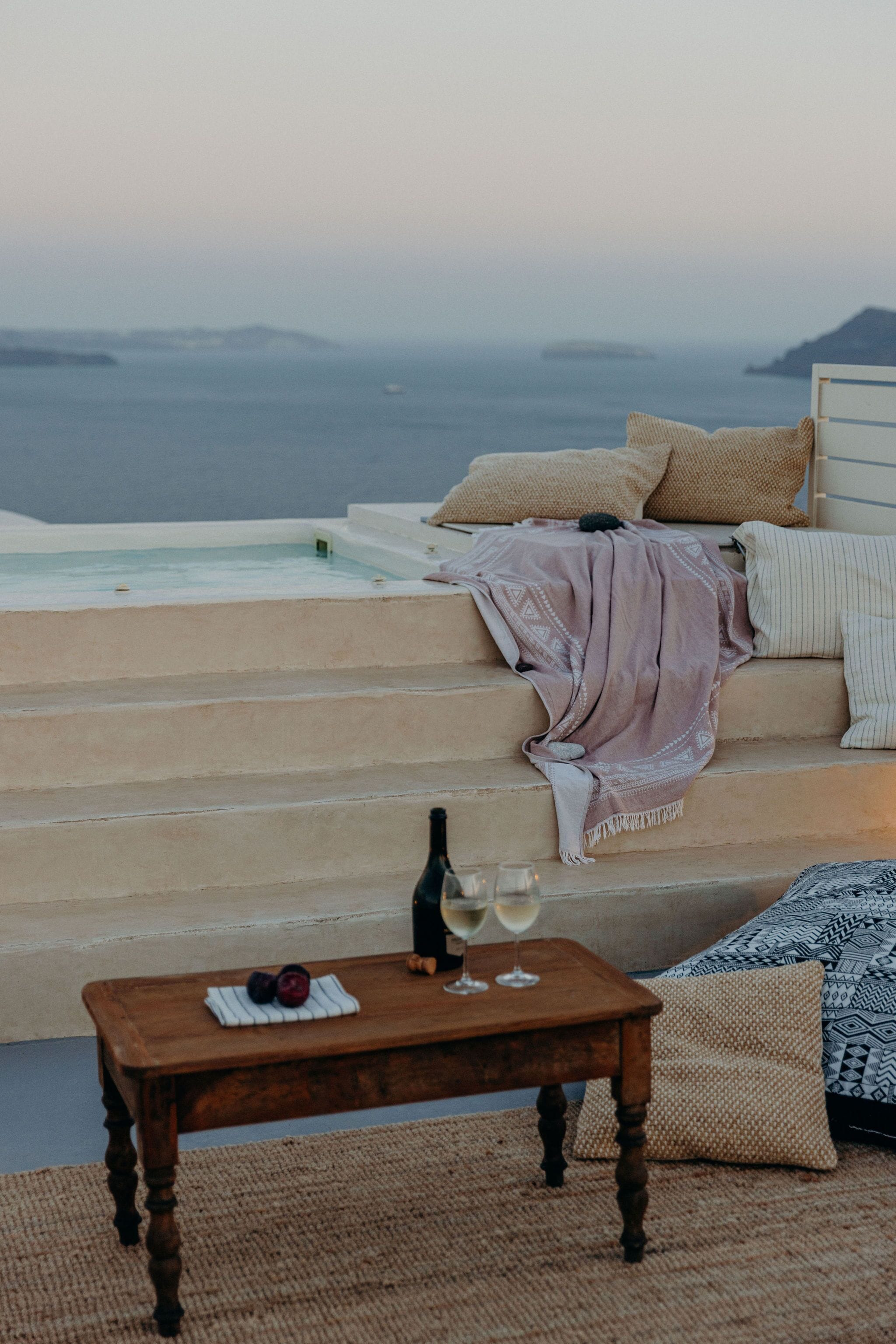 A cozy seating area with a view of the sea, a wooden table with a bottle and glasses, and a patterned cushion.