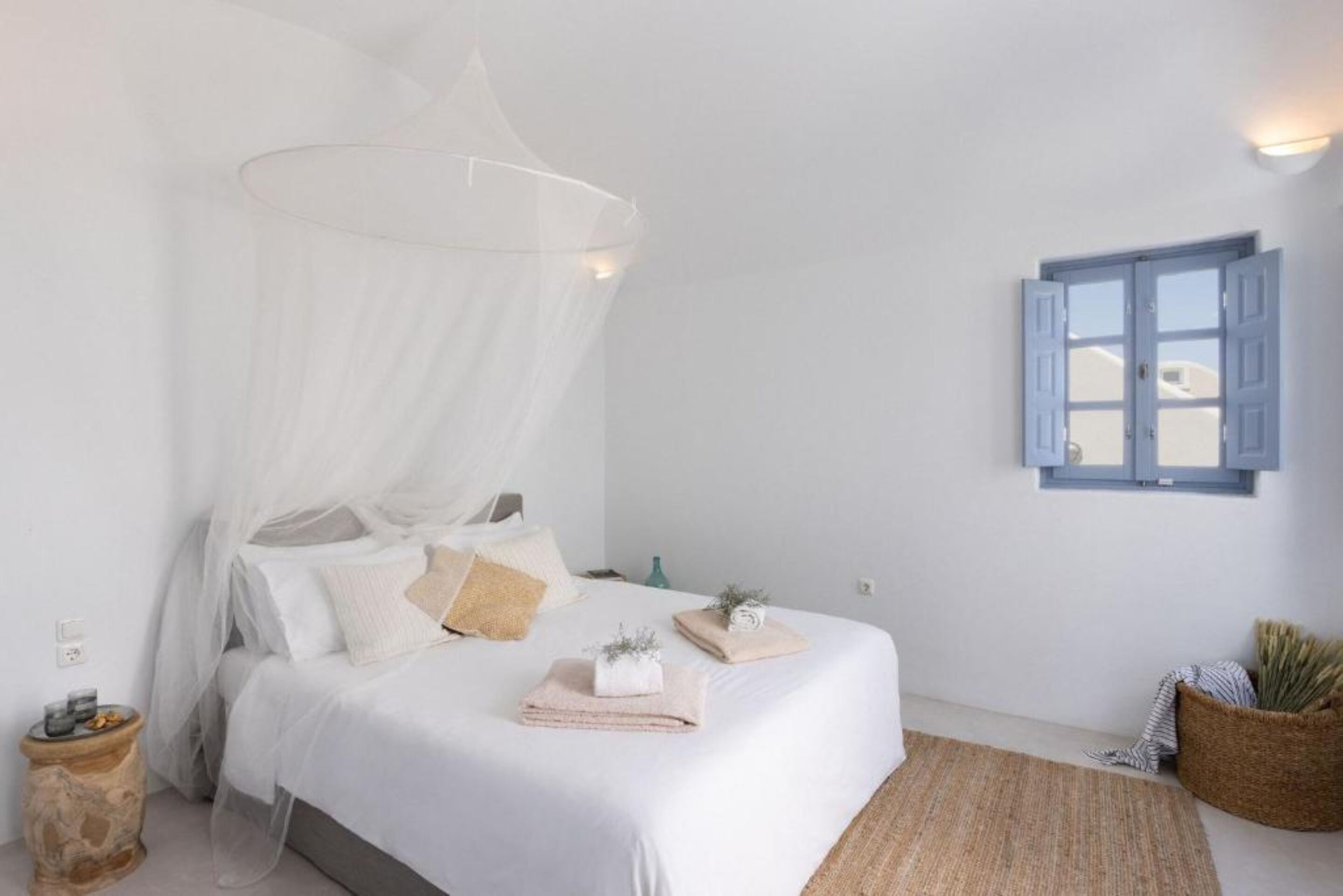 Un dormitorio con cama, ropa de cama blanca, dosel, ventana con contraventanas azules y una cesta tejida en el suelo.