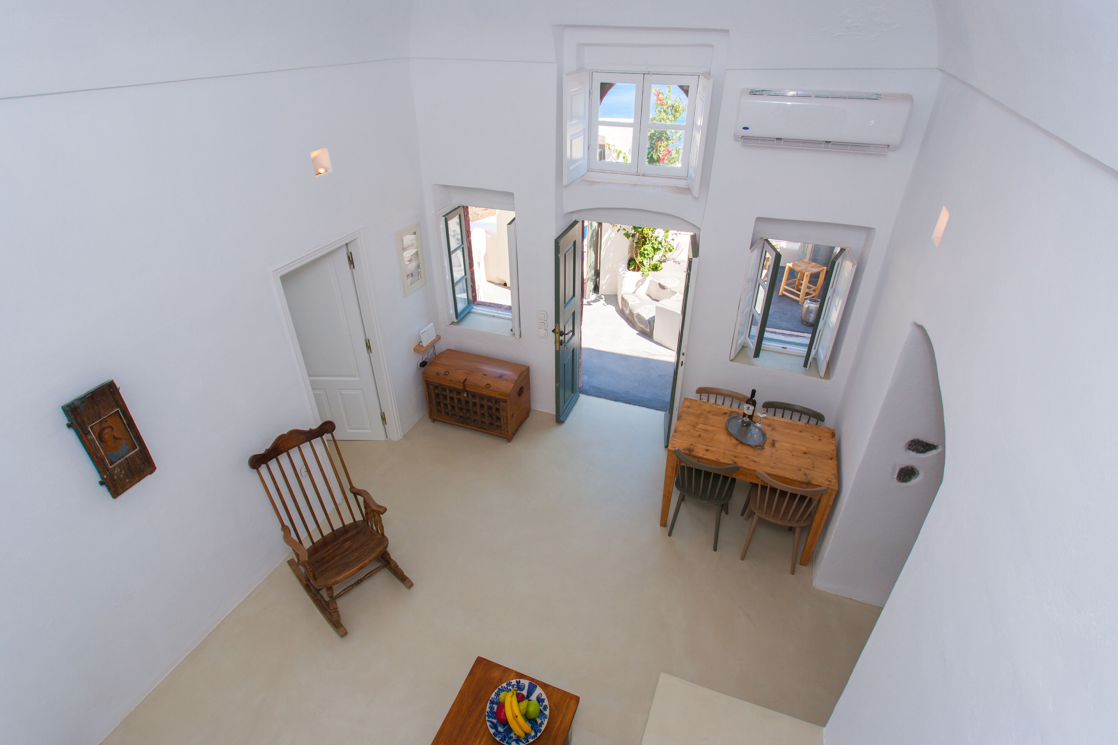 Una habitación amplia con una mesa de comedor de madera, una mecedora y una pequeña mesa con una bandeja de frutas. Hay dos ventanas que ofrecen luz natural y vistas al exterior.