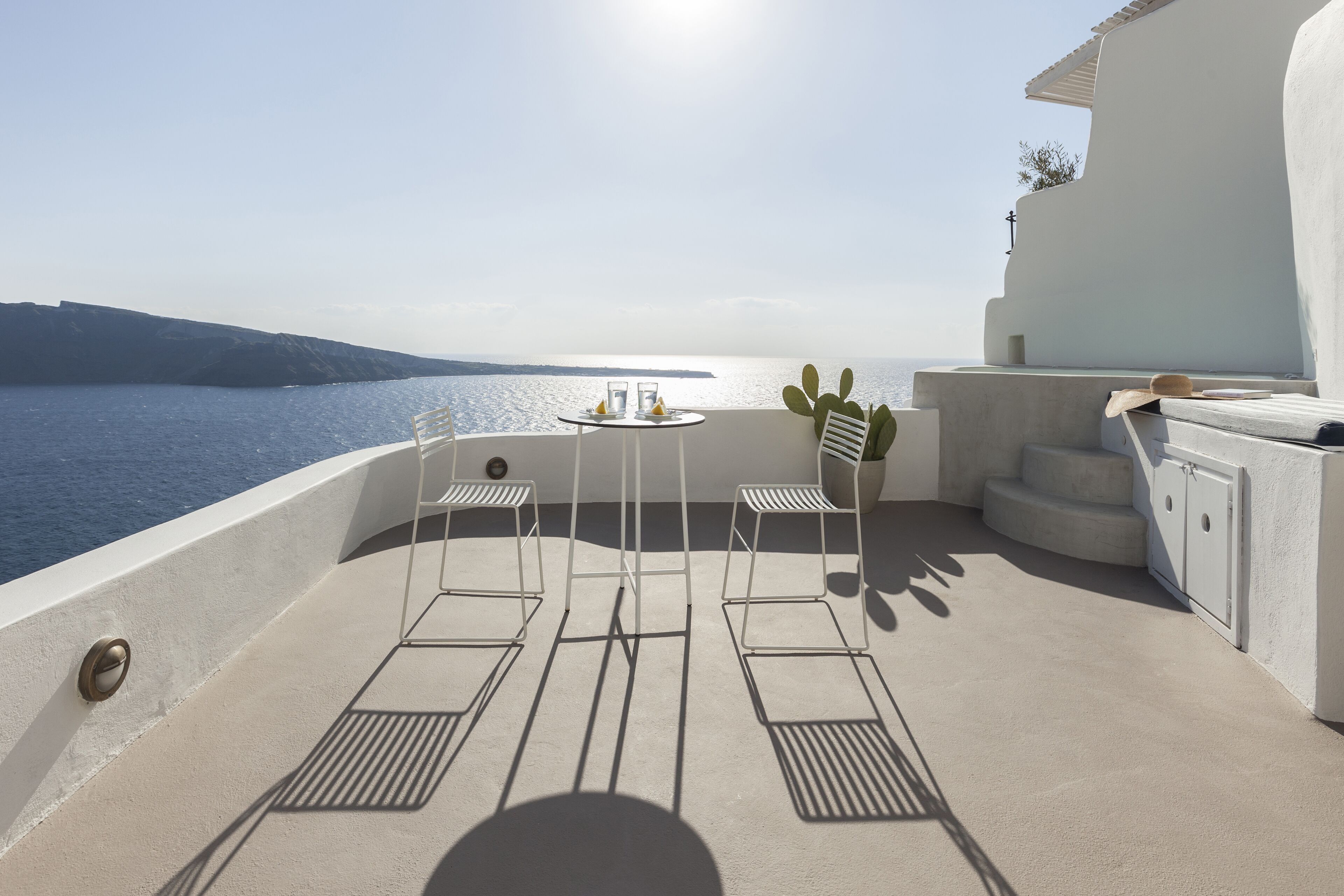 A sunlit terrace with a table and chairs, a potted plant, and a view of the sea.