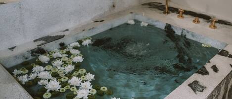 Ein Wellnessbereich mit einer großen Badewanne, gefüllt mit Wasser und Seerosen, einem kleinen Waschbecken und einem Handtuchhalter.