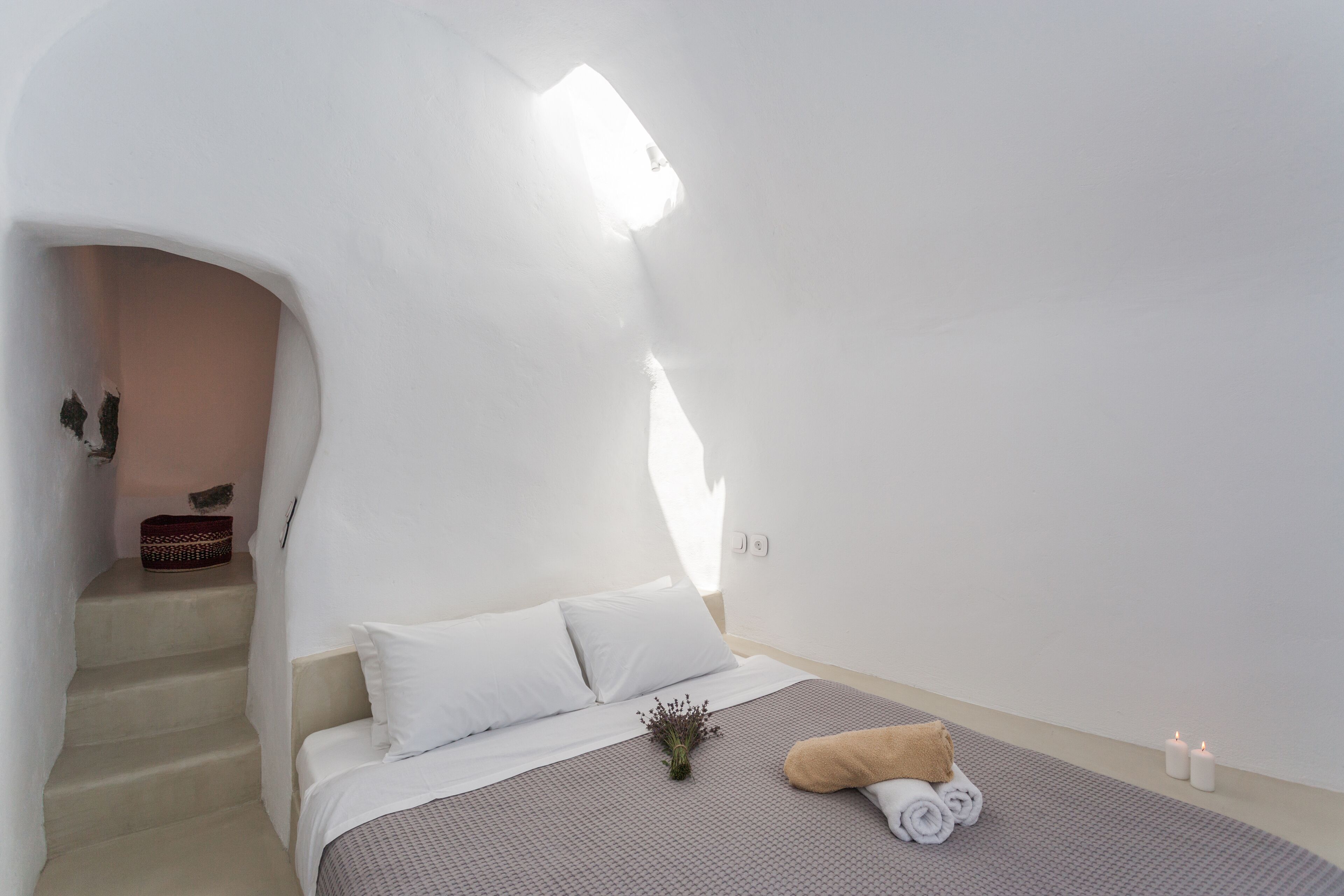 Un dormitorio con cama, almohadas, una toalla enrollada, una planta pequeña y una vela. Se ve una escalera que conduce a otra habitación.