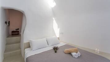 Un dormitorio con cama, almohadas, una toalla enrollada, una planta pequeña y una vela. Se ve una escalera que conduce a otra habitación.