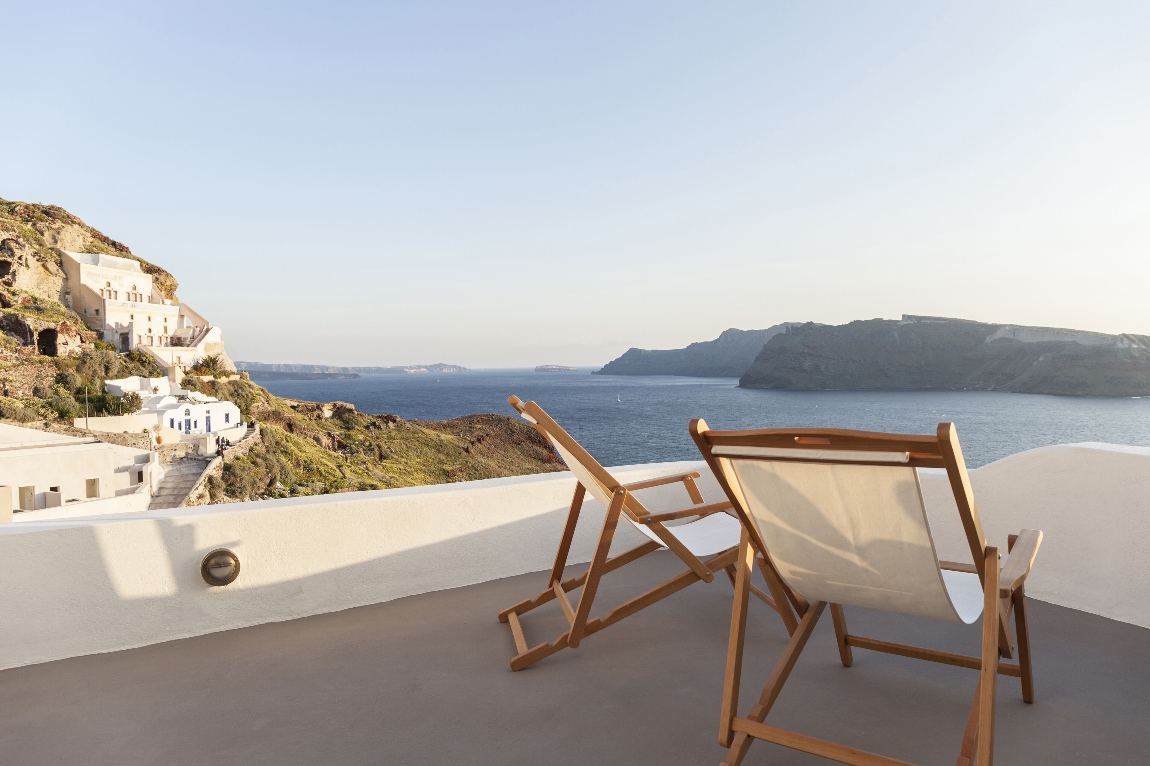A balcony with a wooden chair and a view of a coastal town with white buildings and cliffs.