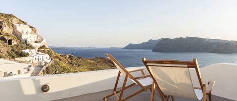 A balcony with a wooden chair and a view of a coastal town with white buildings and cliffs.