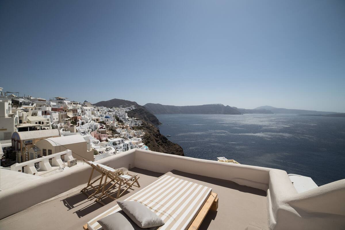 A rooftop terrace with a bed, lounge chairs, and a view of the sea and white buildings.