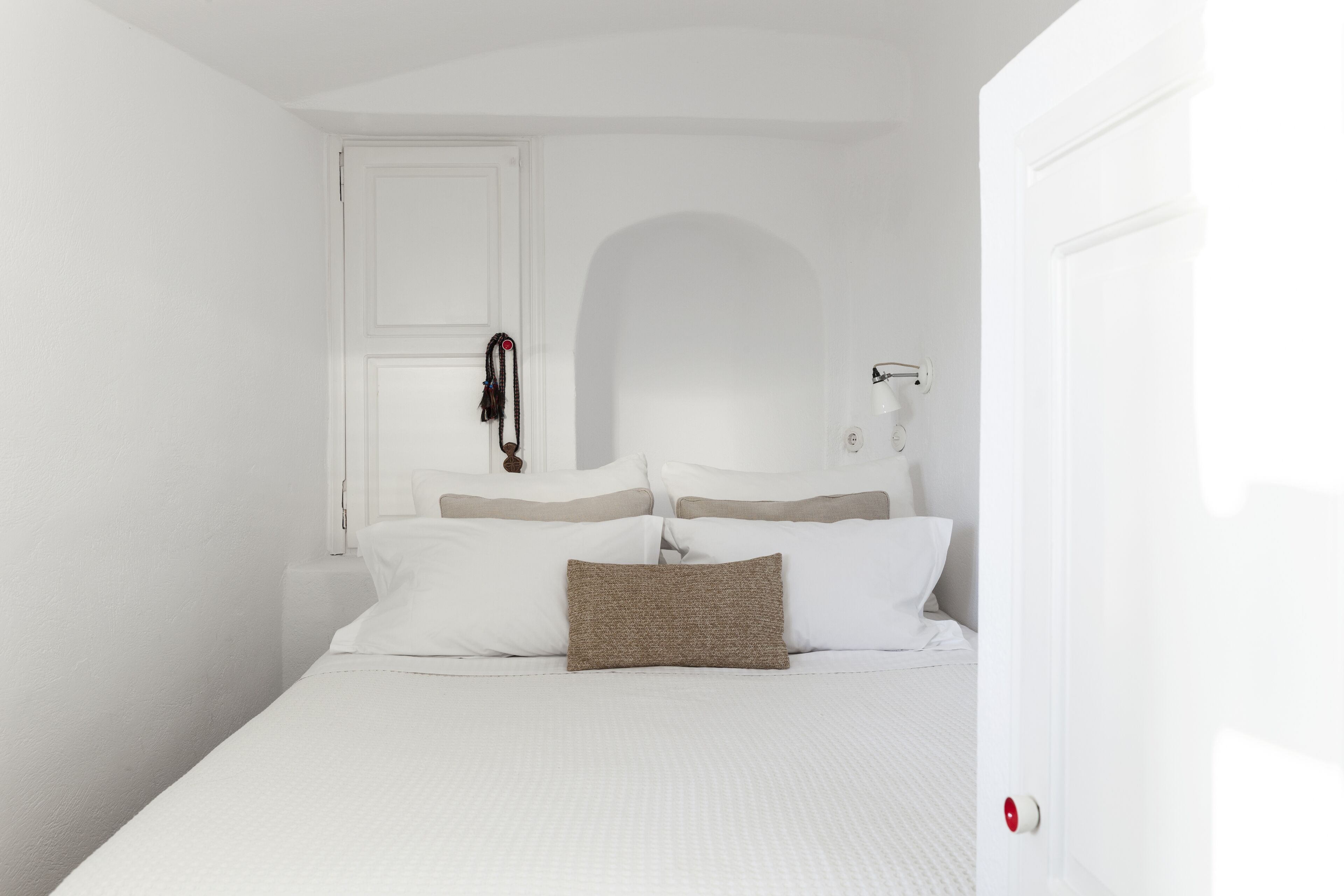 A white room with a bed, a door, and a wall-mounted lamp.