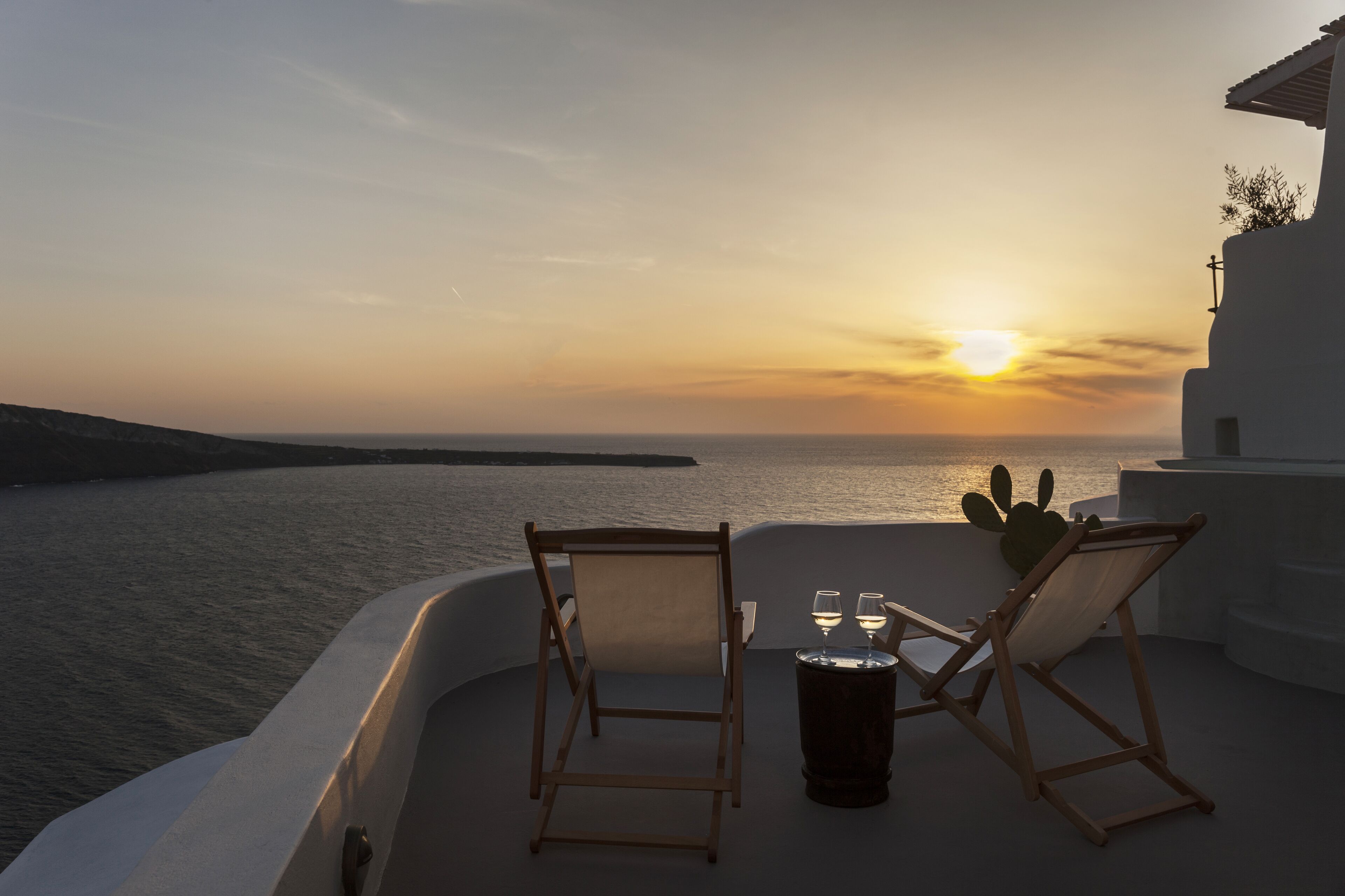 A sunset view from a balcony with two wooden chairs and a table with glasses.