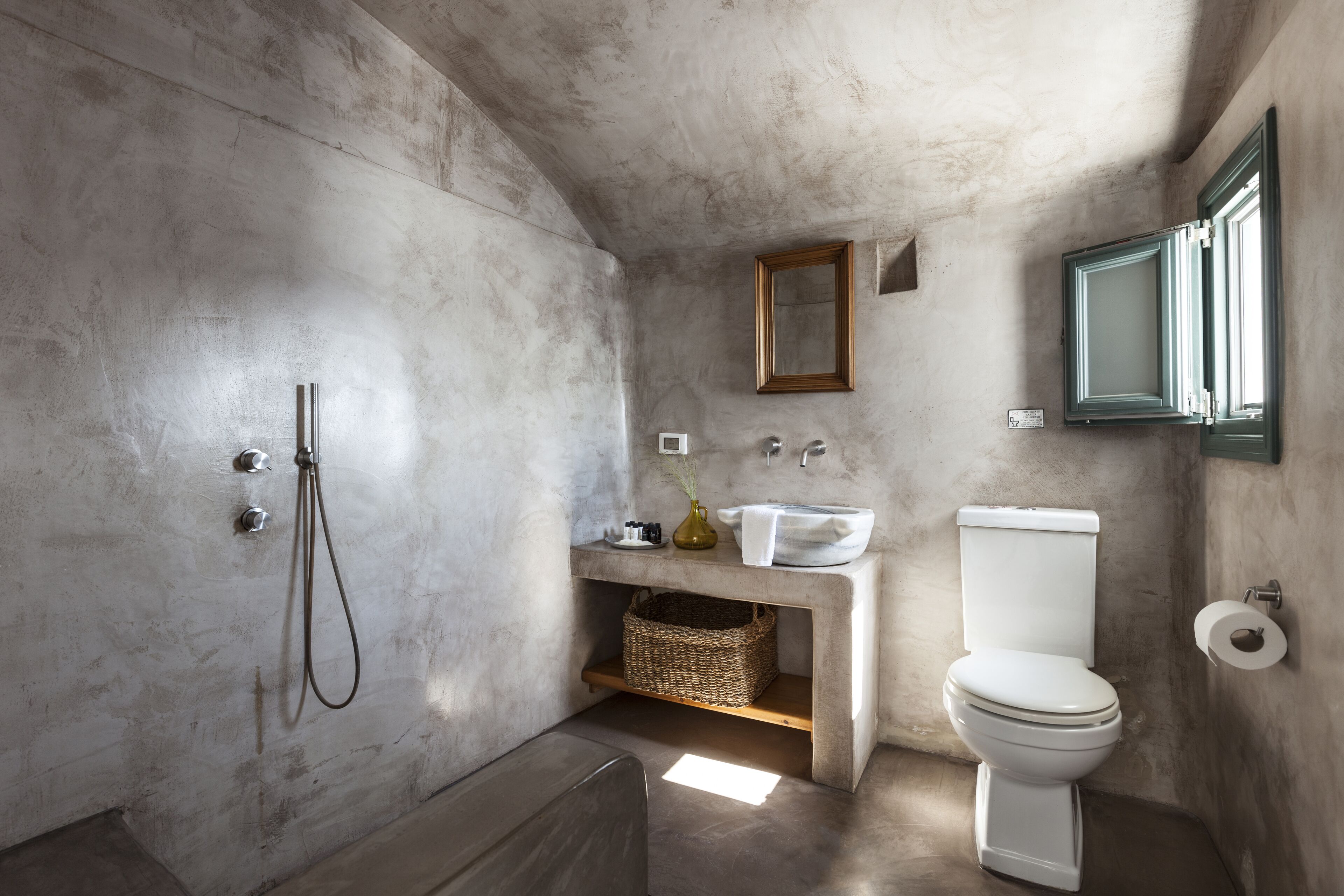 A bathroom with a concrete wall, a shower, a sink, a toilet, and a mirror.