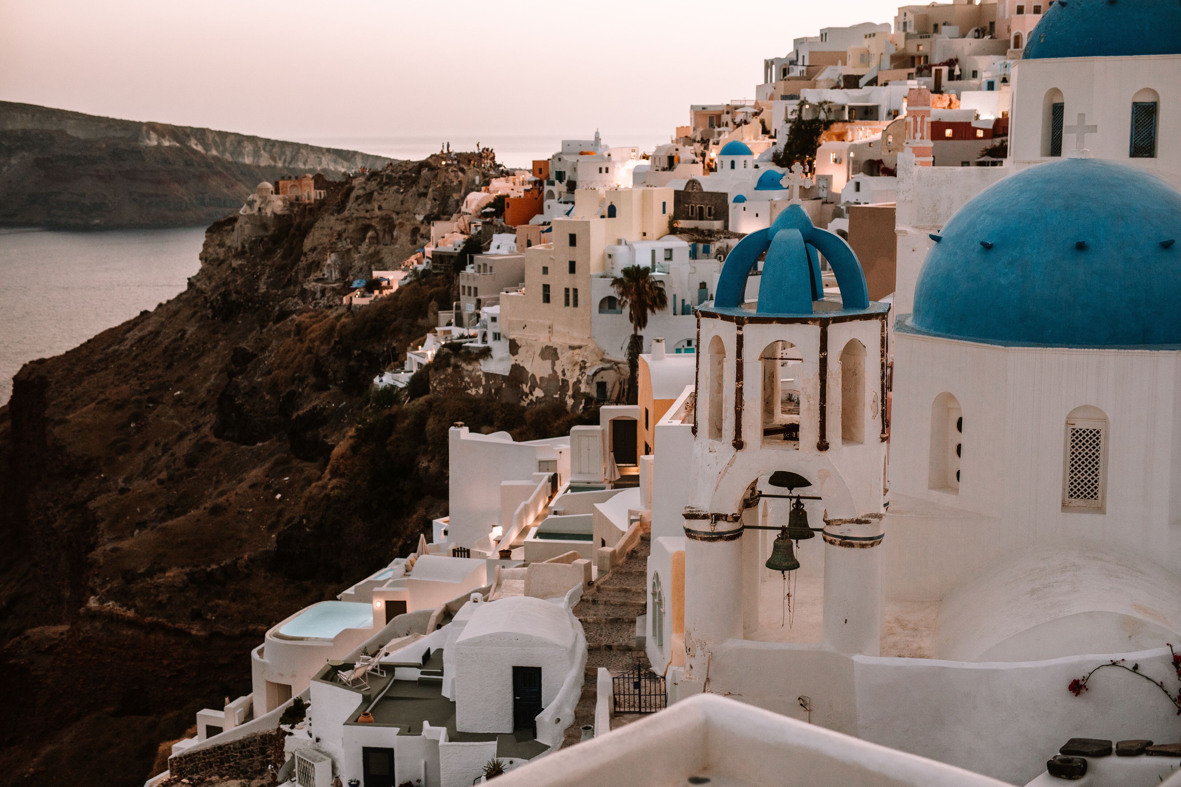 A coastal village with white buildings and blue domes, featuring a rocky cliff and a bridge in the background.