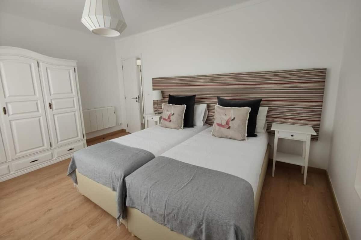 A double bed with a striped headboard, two bedside tables, a white wardrobe, and a hanging light fixture.