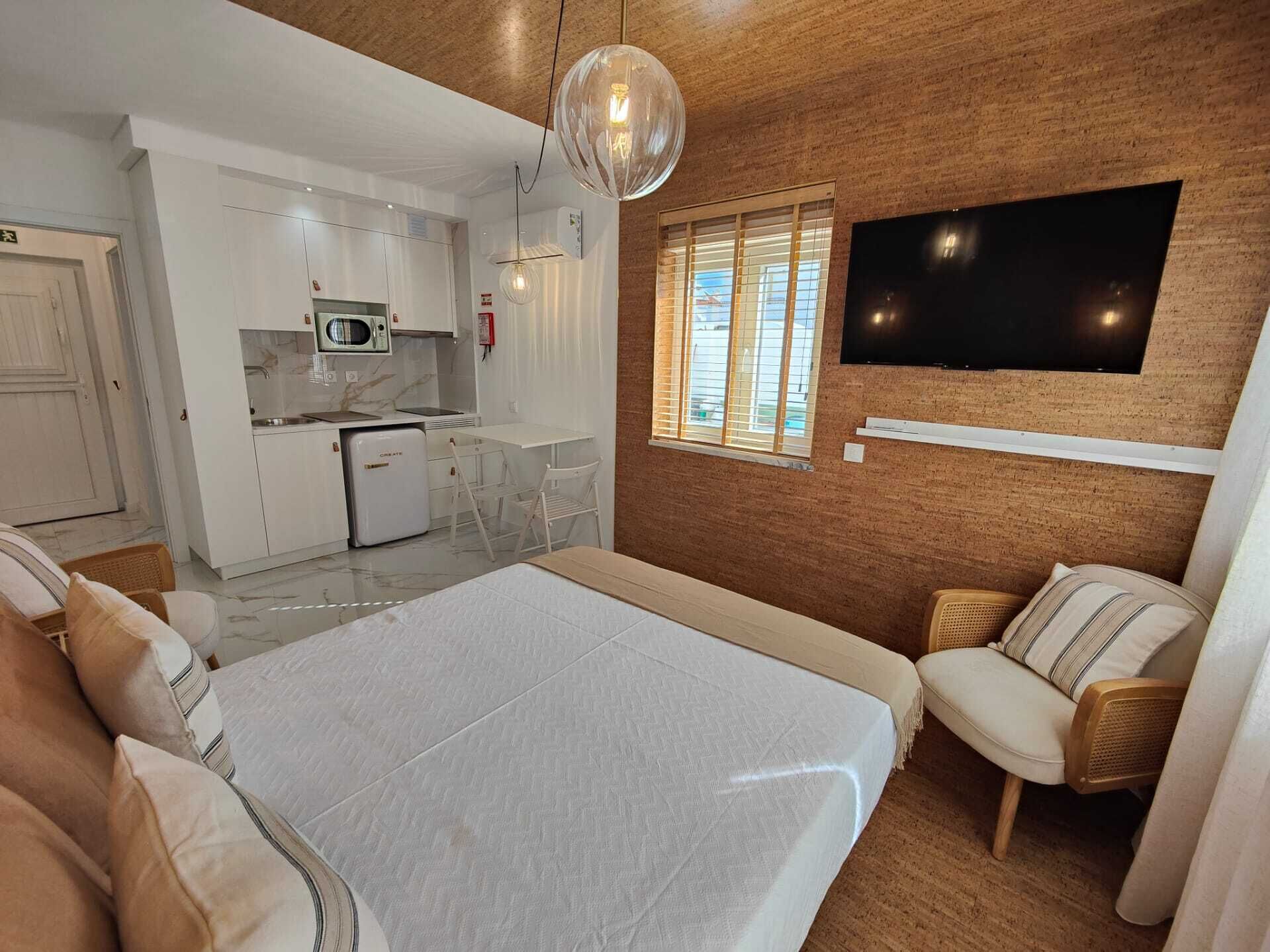 A compact hotel room with a bed, a seating area with a chair and a sofa, a small kitchenette, a TV mounted on the wall, and a window with blinds.