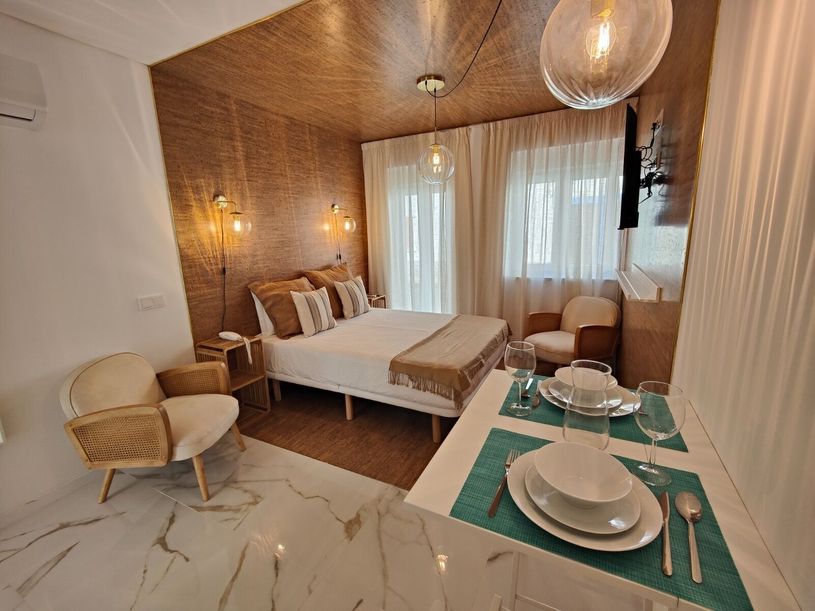 Habitació d'hotel moderna amb un llit gran, una taula de dinar preparada per a dues persones i una paret decorativa de fusta.