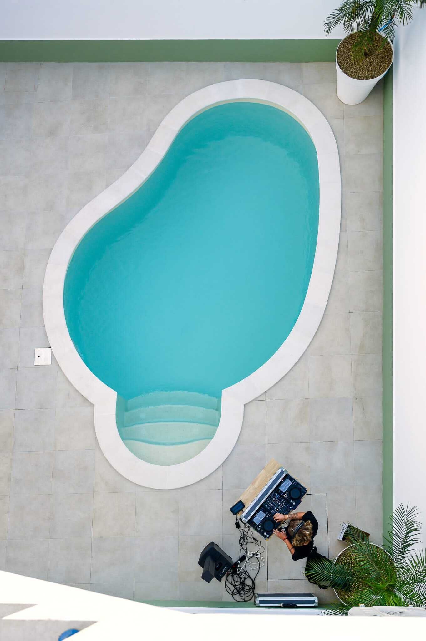 Una piscina de forma peculiar con borde blanco, rodeada por un patio empedrado.