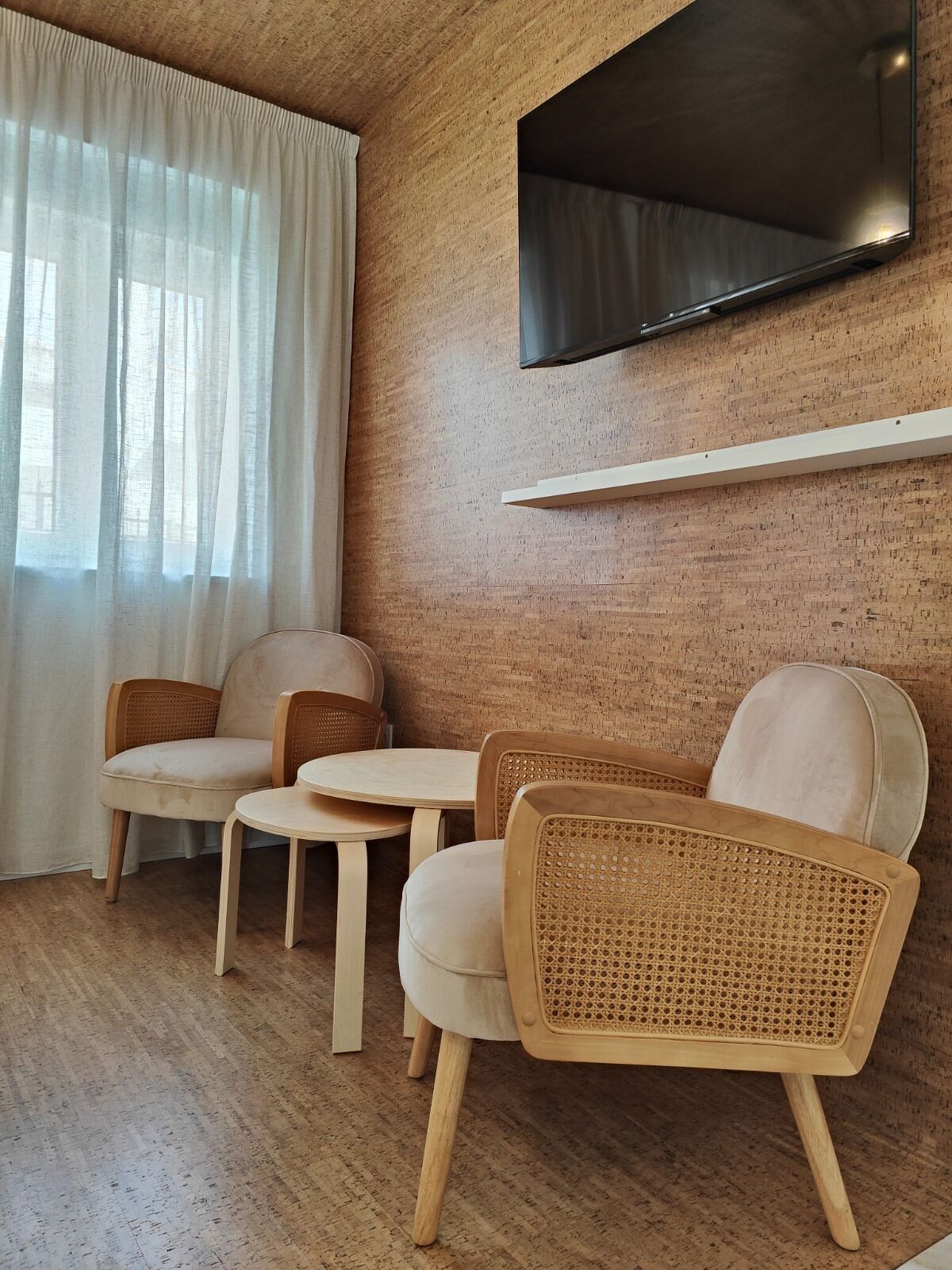 Een kamer met een houten wand, een flatscreen televisie bevestigd boven een wit schap, en een zithoek met twee stoelen en een klein tafeltje.