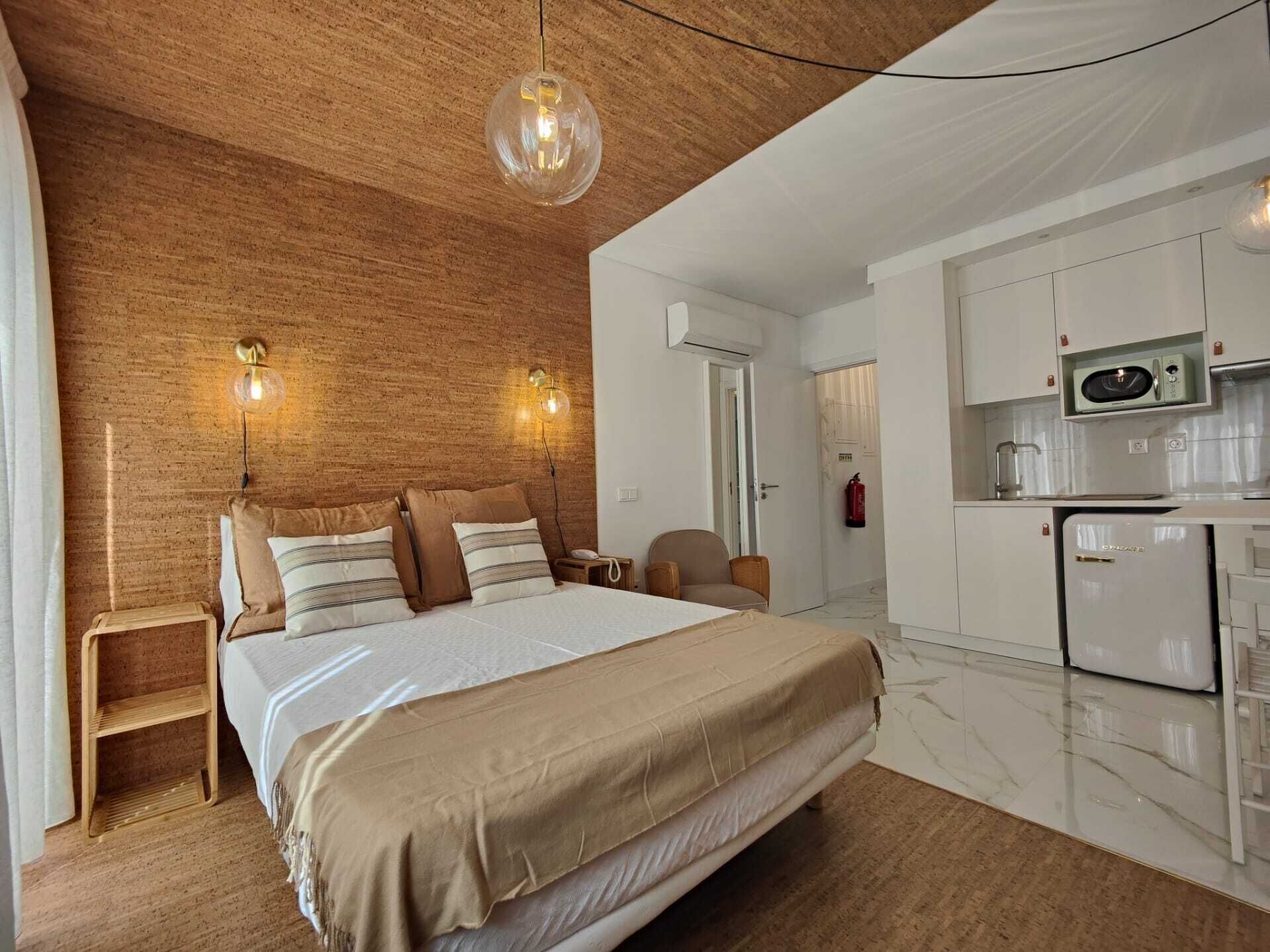 A bedroom with a large bed, a wooden headboard, and a small bedside table. There is a kitchenette with a microwave and a refrigerator.