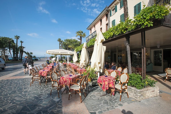 Un café en plein air avec des tables et des chaises, une enseigne indiquant « Caffè Grande Italia », et une zone pavée de pierres.
