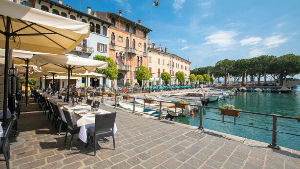 Un patio lungo il fiume con posti a sedere all'aperto, barche ormeggiate nelle vicinanze e edifici storici sullo sfondo.