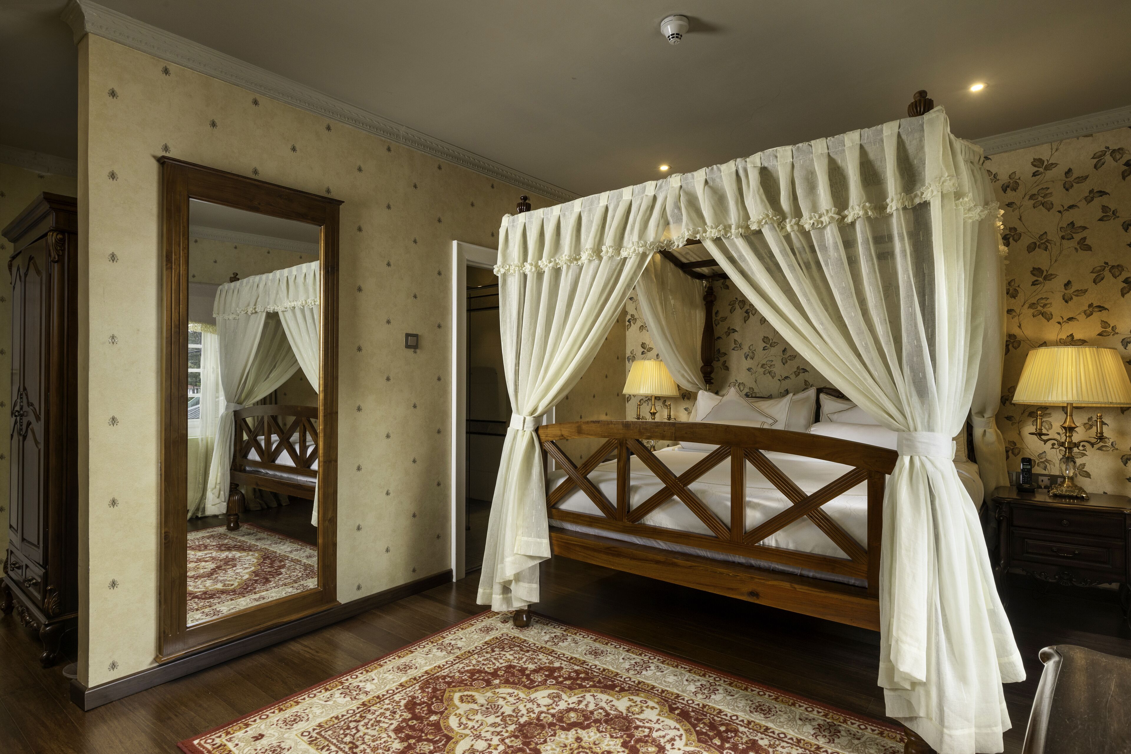 A four-poster bed with a canopy, a large mirror, a nightstand with a lamp, and a patterned rug.