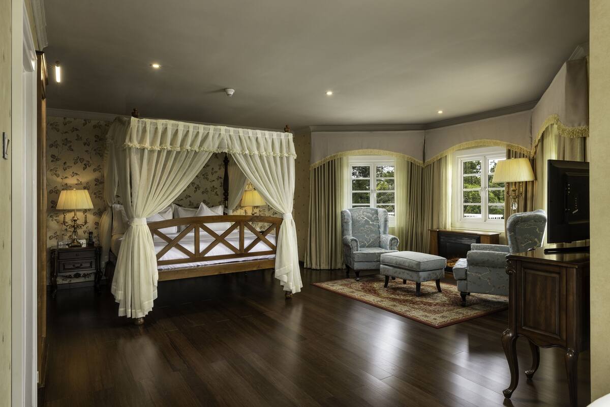 Ein Schlafzimmer mit einem Himmelbett, einer Sitzecke mit Sessel und Hocker, einem Fernseher und einem Kamin.