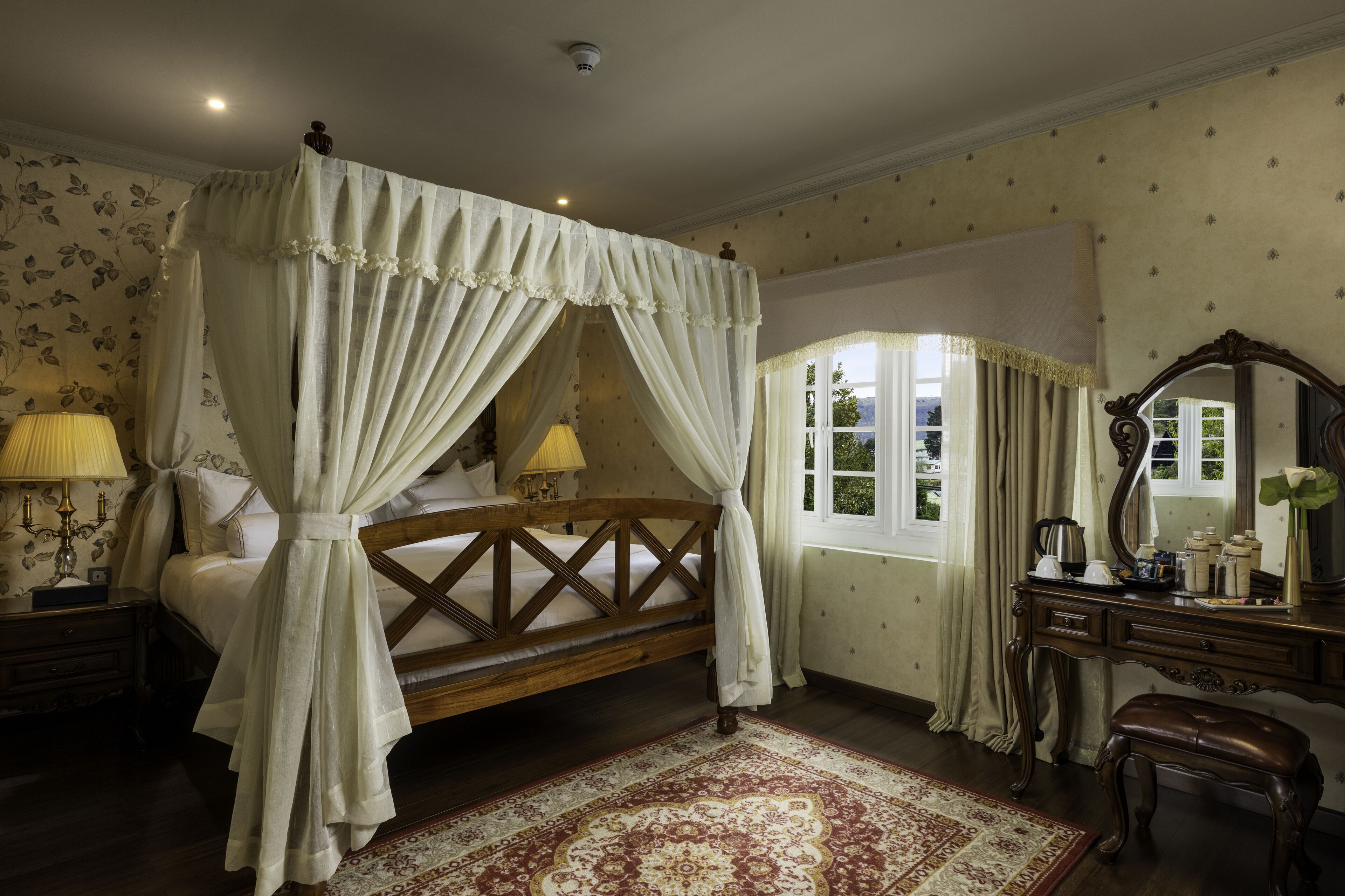 A four-poster canopy bed with a wooden frame, a nightstand with a lamp, a dresser with a mirror, and a window with curtains.