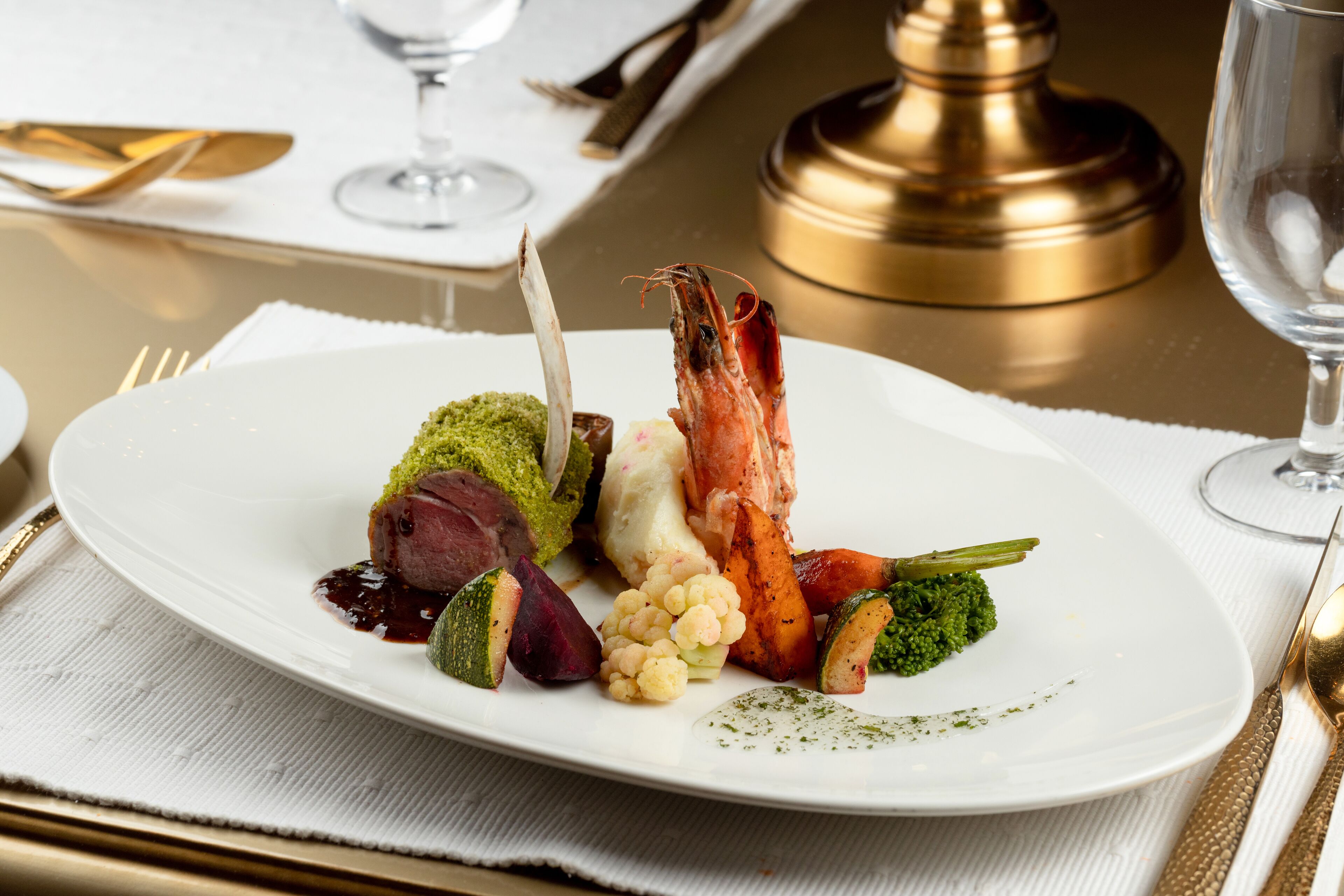 A white plate with a variety of food including a shrimp, vegetables, and a piece of meat, set on a table with a gold-colored salt shaker and a glass of water.