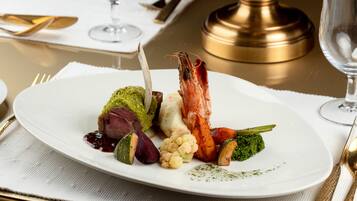 A white plate with a variety of food including a shrimp, vegetables, and a piece of meat, set on a table with a gold-colored salt shaker and a glass of water.