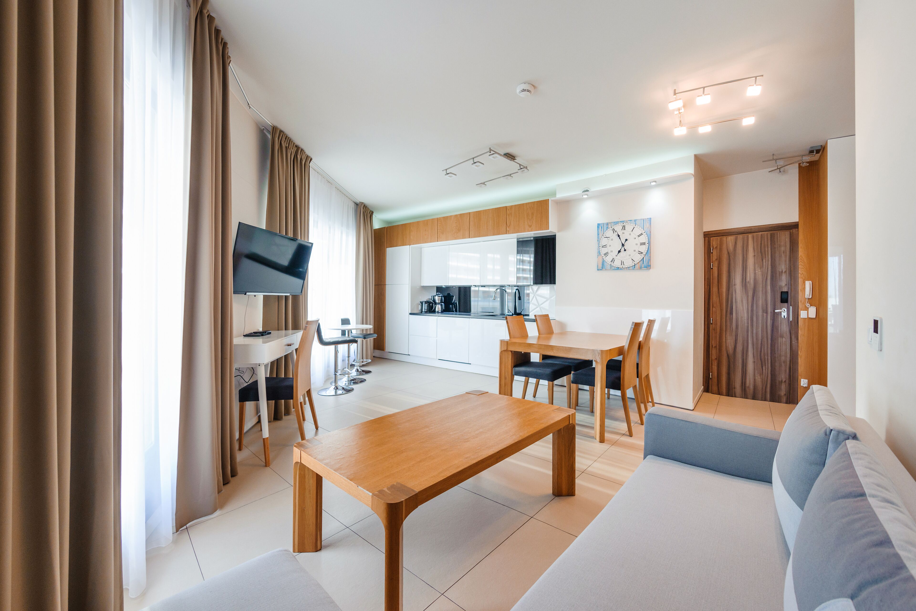 Een moderne woonkamer met een houten salontafel, een bank en een tv op een meubel. Achtergrond is een keuken met een eettafel en stoelen.