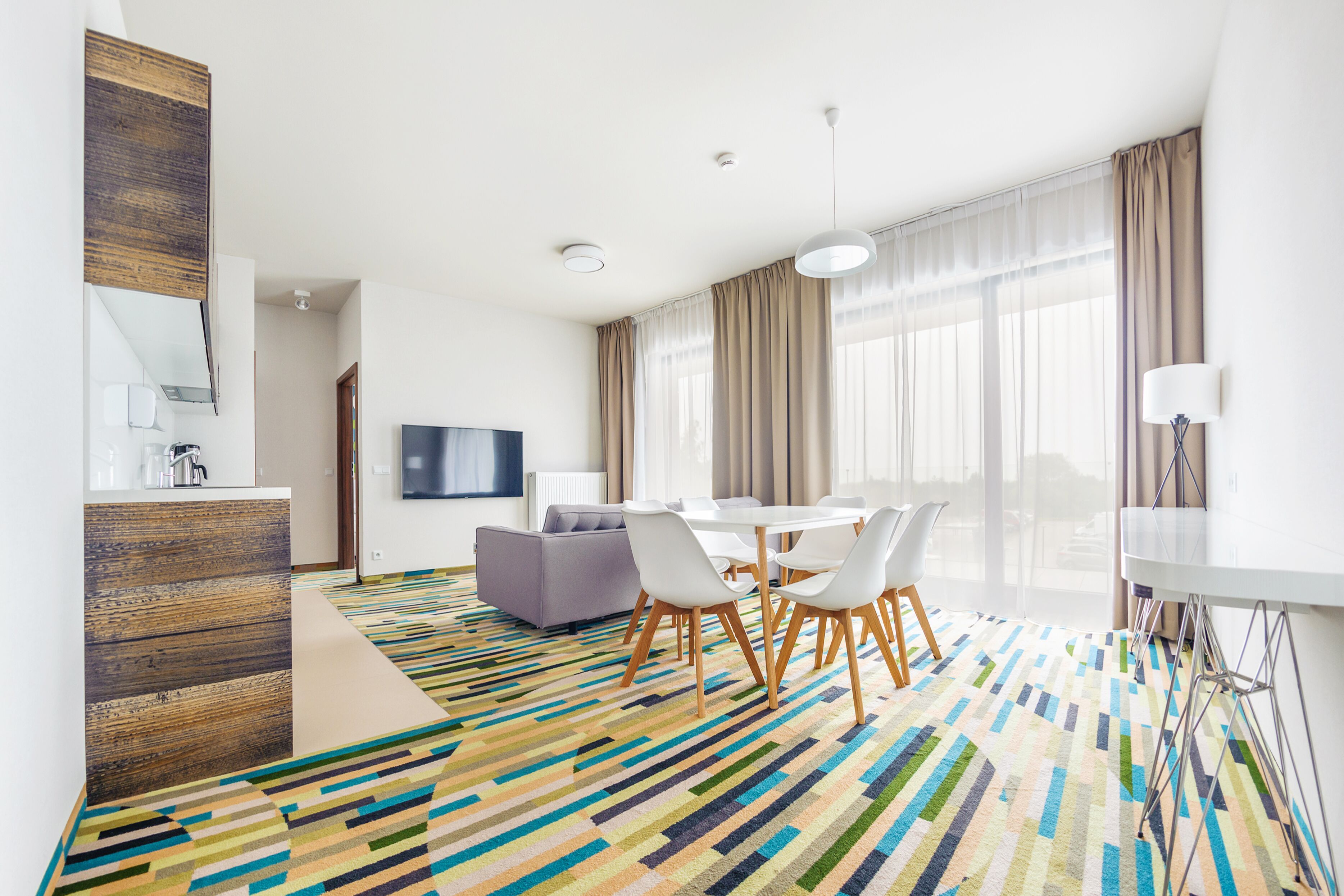 A modern hotel room with a dining area, a sofa, a TV, and a patterned carpet.