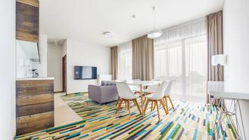 A modern hotel room with a dining area, a sofa, a TV, and a patterned carpet.