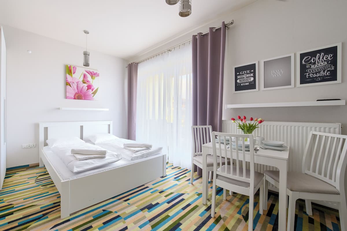 A modern hotel room with a white bed, a colorful striped carpet, a dining area with white chairs, and a window with sheer curtains.