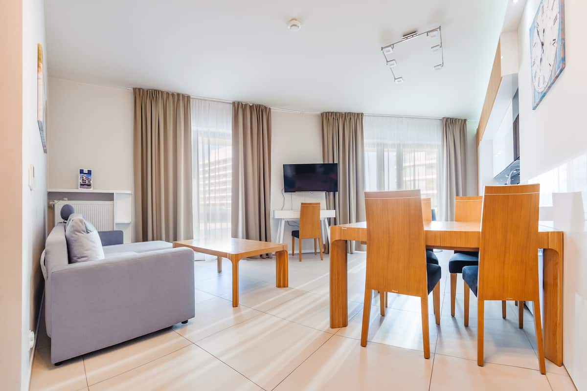 A modern hotel room with a sofa, dining table, chairs, a flat-screen TV, and large windows with curtains.