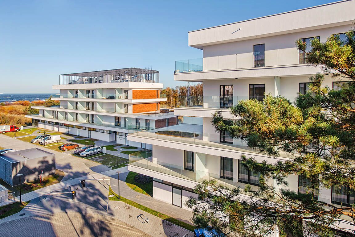 A modern multi-story building with a glass facade, a parking area, and a view of the sea.