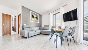 Una sala moderna con un sofá seccional gris, una mesa de comedor de vidrio con sillas, un televisor de pantalla plana montado en la pared y un ventanal que proporciona luz natural.