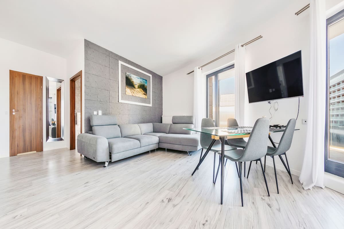 A modern living room with a grey sectional sofa, a glass dining table with chairs, a flat-screen TV mounted on the wall, and a large window providing natural light.