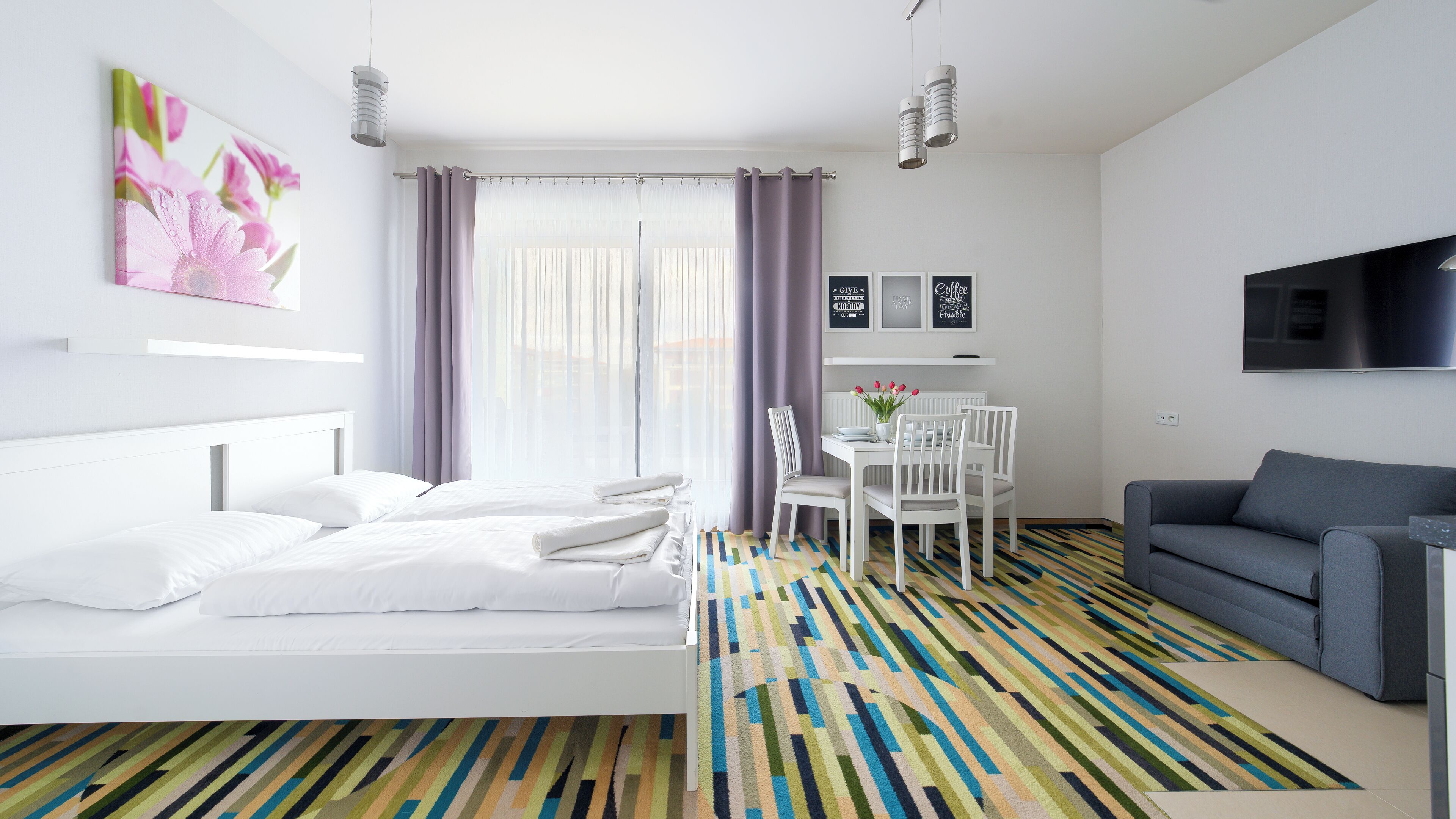 Una habitación de hotel moderna con una cama, un área de comedor con mesa y sillas, un sofá gris y un televisor en la pared.