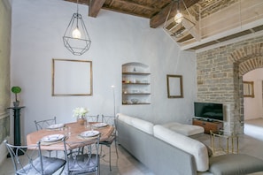Un comedor con una mesa de madera dispuesta para cuatro personas, rodeada de sillas grises tapizadas. Se ve un sofá blanco y un televisor al fondo.
