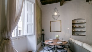 A dining area with a round table, chairs, a hanging light fixture, a sofa, and a shelf with decorative items.